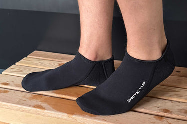 Nahaufnahme von zwei Füßen in schwarzen Neoprensocken auf einem Holzpodest. Ideal für Eisbäder, Kältetherapie und Outdoor-Kälteanwendungen.